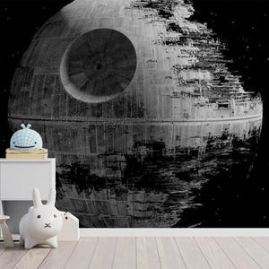 May include: A wall mural featuring a large, detailed black and white depiction of the Death Star in space. The mural is positioned behind a white storage unit, a small white chair, and several small white and blue decorative animal figurines.