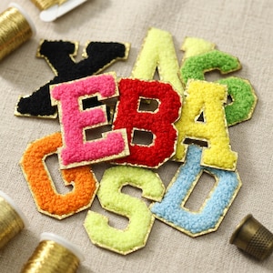 May include: A collection of multi-colored, fuzzy, chenille-style alphabet letter patches with gold-colored metallic borders. The letters are arranged in a cluster and feature vibrant shades of pink, red, orange, yellow, green, blue, and black.
