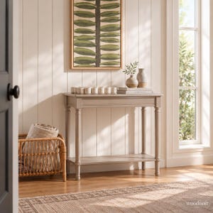 May include: A light gray console table with four turned legs and a lower shelf. On top sits a tray with small cylindrical containers, two ceramic vases, and stacked books. Beside the table is a woven rattan basket holding a folded beige blanket.