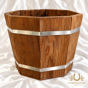 May include: A medium brown wooden planter with a hexagonal shape and a visible wood grain texture. The planter features two horizontal silver-colored metal bands encircling the exterior, positioned near the top and bottom edges of the structure.