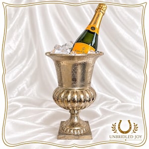 May include: A metallic gold-toned urn-shaped ice bucket with a textured finish, containing clear ice cubes and a bottle of Veuve Clicquot champagne with a yellow label. The vessel features a wide rim, a ribbed bulbous body, and a square base.