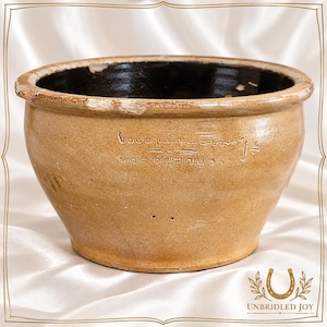 May include: A tan stoneware bowl with a dark brown interior glaze and a thick, rounded rim. The exterior features a stamped inscription with text that appears to be in a stylized script. The bowl has a wide opening and a tapered base.