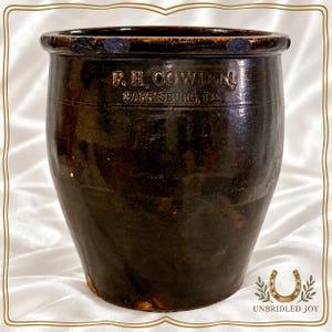 May include: A dark brown glazed stoneware crock featuring a thick rim and a tapered body. The front surface is impressed with the text F.H. COWDEN HARRISBURG, PA. The vessel has a smooth, reflective finish with subtle variations in the dark glaze.