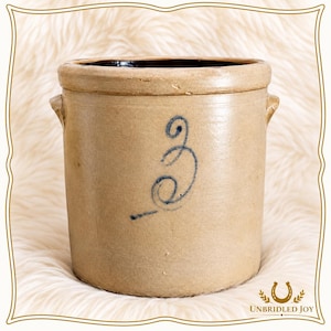 May include: A cylindrical tan stoneware crock featuring a dark blue hand-painted number three on the front. The vessel has a thick rim, two small side handles, and a smooth, glazed finish. The interior of the crock is dark brown in color.