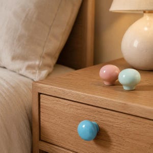 May include: Three round, smooth, glossy cabinet knobs in pastel colors. One bright blue knob is attached to the front of a wooden drawer, while one light pink knob and one light mint green knob are resting on the top surface of the wooden nightstand.