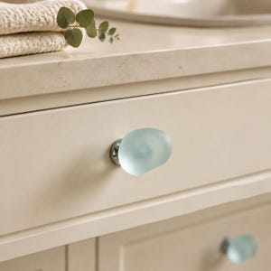 May include: A cabinet knob featuring a smooth, rounded, frosted light blue glass top attached to a polished silver metal base. The knob is mounted on a flat, cream-coloured drawer front, with a second identical knob visible on the lower drawer.