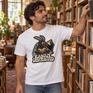 May include: A white short-sleeved t-shirt featuring a graphic of a black moth with red eyes sitting and reading a book under a desk lamp. Below the illustration, the text reads EASILY DISTRACTED in a bold, stylized, cream-colored font.