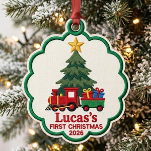 May include: A scalloped-edge fabric Christmas ornament featuring an embroidered green pine tree with a gold star, a red train engine, and a green train car with colorful gifts. The text reads Lucas's FIRST CHRISTMAS 2026 in red embroidery.
