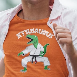 May include: An orange t-shirt featuring a graphic of a green dinosaur wearing a white martial arts uniform with a black belt. The text JITSUSAURUS is printed in white above the dinosaur. The shirt is worn under a light pink button-down shirt.