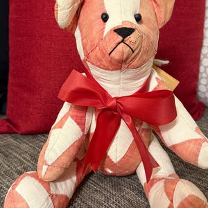 May include: A stuffed teddy bear constructed from a patchwork of cream and coral-colored fabric panels. The bear features small black bead eyes, a stitched black nose, and a vibrant red satin ribbon tied in a bow around its neck.