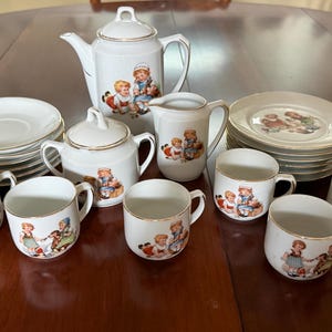 May include: A white porcelain tea set featuring gold trim and colorful illustrations of children playing. The set includes a teapot with a lid, a sugar bowl with a lid, a creamer, six cups, and six saucers, all decorated with matching vintage-style motifs.