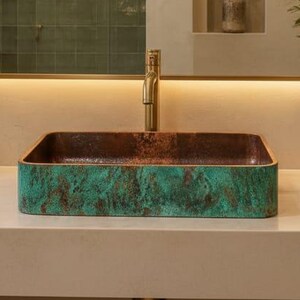 Op de afbeelding: Een rechthoekige waskom met een verweerde koperen en groen-patina afwerking, geplaatst op een glad beige aanrechtblad. De waskom is voorzien van een goudkleurige kraan en bevindt zich onder een rechthoekige spiegel met geïntegreerde verlichting.