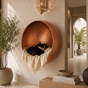 Puede incluir: Una cama para gatos circular montada en la pared de cobre con textura martillada. Dentro de la estructura en forma de cuenco, un gato blanco y negro descansa sobre una manta de tela texturizada de color crema. El metal refleja la luz.