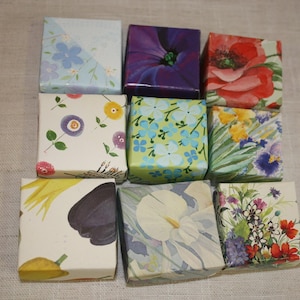 May include: A collection of nine square boxes, each wrapped in paper featuring distinct floral and botanical patterns. The designs include red poppies, purple flowers, blue blossoms, yellow accents, and a white calla lily, arranged in a grid.
