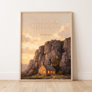 May include: A framed wall art print featuring a landscape painting of a small stone cottage nestled at the base of a large, rugged cliff under a sunset sky. The text GOD IS OUR REFUGE AND STRENGTH PSALM 46:1 is printed in white at the top centre.