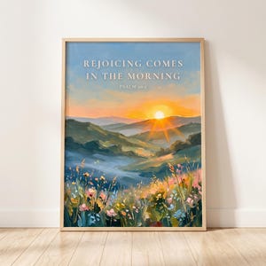 May include: A framed wall art print featuring an oil painting of a sunrise over rolling hills and a wildflower meadow. The text REJOICING COMES IN THE MORNING PSALM 30:5 is printed in white at the top centre against a blue and orange sky.