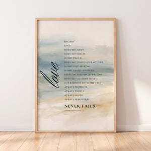 May include: A framed vertical wall art print featuring a watercolour background in shades of blue, beige, and white. The text reads: Love is patient, kind, does not envy, does not boast, is not proud, does not dishonour others, never fails, 1 Corinthians 13.