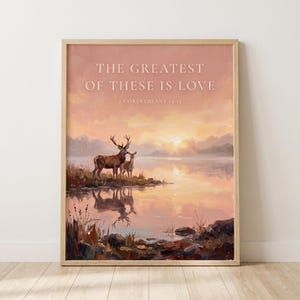 May include: A framed wall art print featuring a sunset landscape with a deer and a fawn standing by a calm lake. The text reads THE GREATEST OF THESE IS LOVE and 1 CORINTHIANS 13:13 in white lettering against a soft pink and orange sky background.