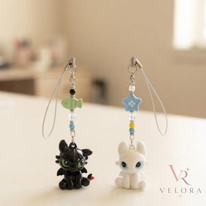 May include: Two phone charms featuring small dragon figures. The left charm has a black dragon with green eyes, a green fish bead, and clear and blue accents. The right charm has a white dragon with blue eyes, a blue star bead, and clear and yellow accents.