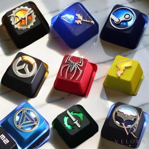 May include: A collection of nine custom mechanical keyboard keycaps featuring various icons and symbols, including a gear, a blue crystal, the Steam logo, a spider emblem, a lightning bolt, and a bat symbol, rendered in diverse colours.