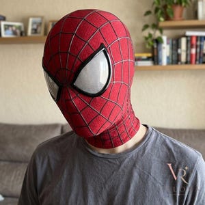 May include: A red fabric mask featuring a black web pattern and large, white, mesh-covered eye lenses. The mask covers the entire head and neck area, displaying a textured surface with a distinct grid design across the front and sides.