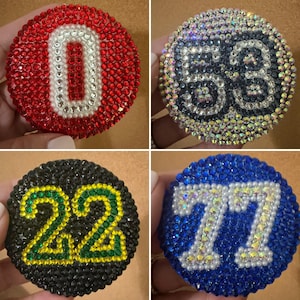 May include: Four circular coasters decorated with small rhinestones. One is red with a white number 0, one is iridescent with a black number 53, one is black with a green and yellow number 22, and one is blue with a white number 77.