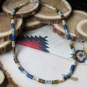 May include: A beaded necklace featuring cylindrical beads in alternating colors of brown tiger eye, cream, bright blue, and dark blue, separated by small silver-toned metal spacers. The necklace is secured with a silver-toned metal lobster claw clasp.