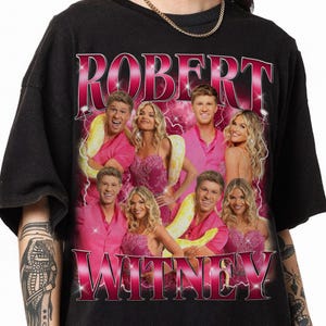 May include: A black short-sleeve t-shirt featuring a graphic print of Robert and Witney in various poses. The design includes pink lightning accents, a pink gradient background, and the text ROBERT at the top and WITNEY at the bottom in bold font.