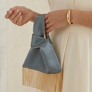Puede incluir: Un bolso de mano de satén azul pizarra con un diseño de asa superior anudada y un largo fleco dorado brillante en el borde inferior. El bolso tiene un acabado de tela suave y estructurado con una forma triangular distintiva y definida.