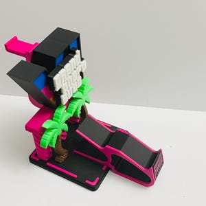 May include: A black and hot pink plastic stand featuring a rectangular base, a ramp-like support, and a vertical structure decorated with two bright green palm trees and a white blocky sign with raised lettering against a dark background.