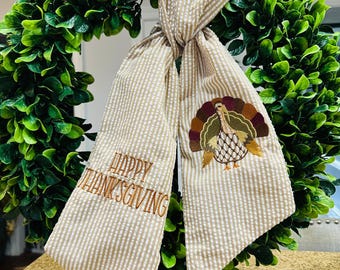 May include: A beige and white seersucker fabric ribbon tied in a knot, featuring the embroidered brown text Happy Thanksgiving and a colorful embroidered turkey with brown, gold, and green feathers on the right side of the fabric.