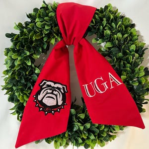 May include: A circular green boxwood wreath adorned with a bright red fabric bow. The left tail of the bow features an embroidered bulldog head with a spiked collar, while the right tail displays the white embroidered text UGA in a serif font.
