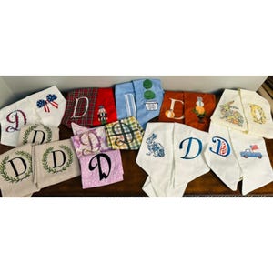 May include: A collection of various fabric towels featuring embroidered designs, including the letter D in multiple fonts, floral patterns, a nutcracker, pumpkins, rabbits, and a blue truck with an American flag, displayed in a group.