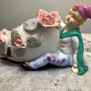 May include: A porcelain figurine of a person wearing a purple hat, white jacket, and light blue pants with red accents, sitting beside a large white shoe. The shoe is decorated with three-dimensional pink roses and green leaves.
