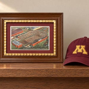 May include: A framed vintage postcard print of Memorial Stadium at the University of Minnesota with a dark wood and gold frame, alongside a maroon baseball cap featuring a gold embroidered M logo and a stack of books with Minnesota spine text.