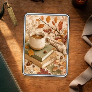 Könnte beinhalten: Ein digitales Tablet mit einer Illustration von einer dampfenden Kaffeetasse auf zwei gestapelten Büchern, umgeben von Herbstblättern, Eicheln und einer gestrickten Stofftextur, alles vor einem warmen, saisonalen und gemütlichen Hintergrund.