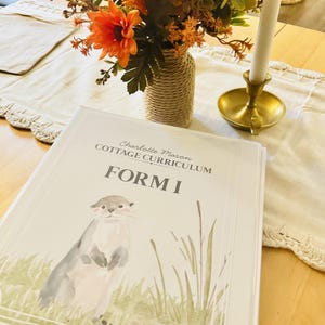 Op de afbeelding: Een witte map met de tekst Charlotte Mason COTTAGE CURRICULUM FORM I op de kaft, voorzien van een aquarelillustratie van een otter die in groen gras bij het water staat, geplaatst op een houten tafel met een bloemstuk.
