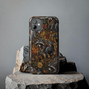 Könnte beinhalten: Eine Smartphone-Hülle mit einem Muster im Buntglas-Stil, das Waschbären inmitten von Herbstblättern, Eicheln und Beeren zeigt. Das Design verwendet eine Palette aus tiefen Braun-, Orange-, Gelb- und Grüntönen mit dunklen Konturen.