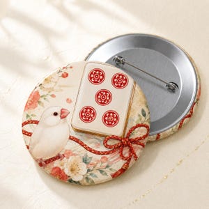 May include: A round pinback button featuring a white Java sparrow perched on a red cord next to a mahjong tile with five red floral pips. The background displays a soft floral pattern with red and white blossoms and a metallic pin mechanism.