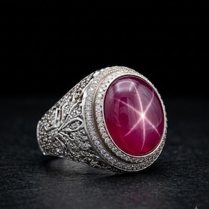 Könnte beinhalten: Ein Silberring mit einem großen, ovalen, tiefroten Sternrubin-Cabochon als Herzstück. Der Stein zeigt einen deutlichen sechsstrahligen Sterneffekt. Der Ring ist mit filigranen Mustern und kleinen weißen Steinen detailliert verziert.