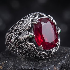 Op de afbeelding: Een zilveren ring met een ovaal geslepen dieprode edelsteen in het midden. De band is voorzien van gedetailleerd metaalwerk met twee adelaarachtige vogels met gespreide vleugels, elk met kleine groene accenten voor de ogen van de vogels.