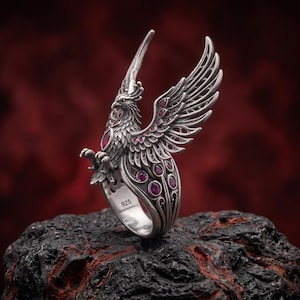 Pode incluir: Um anel de prata esterlina com um design detalhado de fénix com asas abertas. A ave é acentuada com várias pequenas pedras preciosas vermelhas redondas e uma pedra vermelha de corte marquise no peito. A banda interior tem o carimbo 925.
