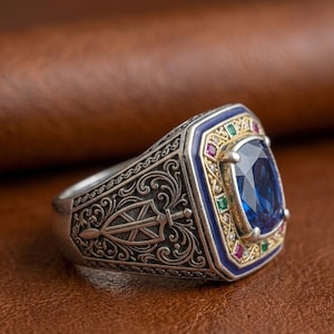 Op de afbeelding: Een zilveren ring met een ovale blauwe middensteen omringd door een goudkleurig frame met kleine groene en rode vierkante accenten. De band is voorzien van gedetailleerde gravures en een schild- en zwaardmotief op het zijprofiel.