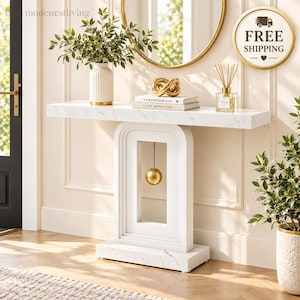 May include: A white console table with a faux marble finish and a central arch-shaped base featuring a suspended gold sphere. The tabletop holds a ribbed white vase with greenery, two stacked books, a gold knot sculpture, and a glass reed diffuser.