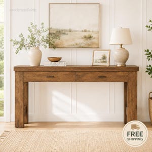 May include: A rectangular wooden console table with two drawers and dark metal pulls. On top sits a beige ceramic vase with green foliage, a small wooden bowl, a stack of books, a framed landscape print, and a table lamp with a white shade.