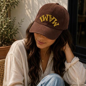May include: A brown corduroy baseball cap featuring the word MAMI embroidered in yellow capital letters across the front panel. The cap has a structured crown, a curved brim, and a button on top, showcasing a classic six-panel design.
