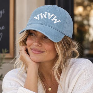 May include: A light blue baseball cap with a curved brim and the word MAMA embroidered in white capital letters across the front. The cap features a distressed, washed-out fabric texture and a structured crown with a button on the top center.