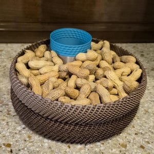 May include: A round, dark brown, textured bowl with a cross-hatched pattern filled with numerous light brown, unshelled peanuts. Centered within the peanuts is a small, cylindrical, bright blue container with a textured, diamond-patterned rim.