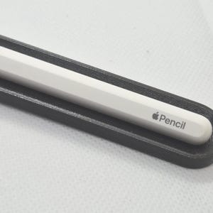 May include: A white Apple Pencil with a smooth, cylindrical body and a rounded tip, resting securely inside a black, elongated, 3D-printed holder with a textured, layered surface finish that follows the contour of the stylus.