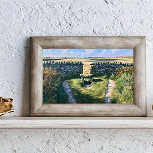May include: A rectangular wooden frame containing a landscape print of a stone wall with a gap, a stone stile, and a grassy path leading through the opening into a field under a blue sky, flanked by a decorative shell and a piece of driftwood.